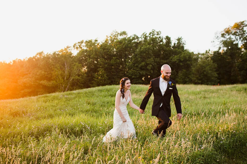 Bride and groom walk across a sunlit hillside meadow at Tower Hill Botanic Garden with the sun flaring behind the trees