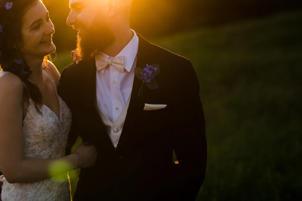 Close portrait of a bride and groom in warm backlight at Tower Hill Botanic Garden, the groom in a black tuxedo with a blue boutonniere