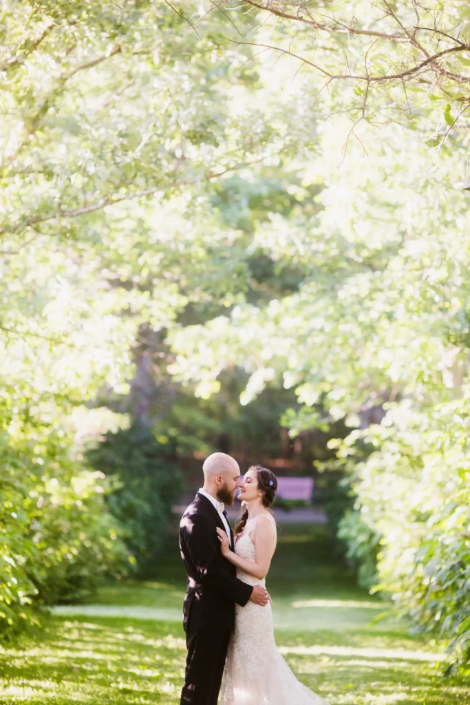 Bride and groom kiss beneath sunlit trees on a garden path at Tower Hill Botanic Garden in Boylston, MA
