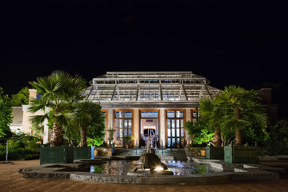 The Orangerie at Tower Hill Botanic Garden lit up at night with palms and the Winter Garden fountain in the foreground