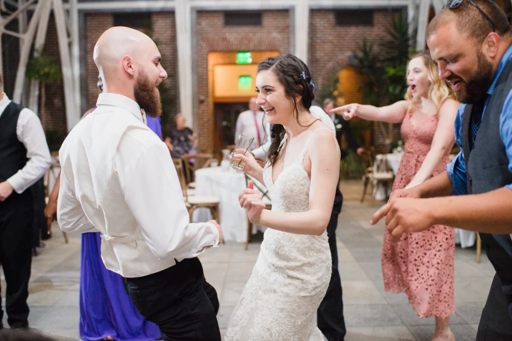 Bride and groom dance with friends on the Orangerie floor at Tower Hill Botanic Garden with the brick walls and courtyard doors behind them