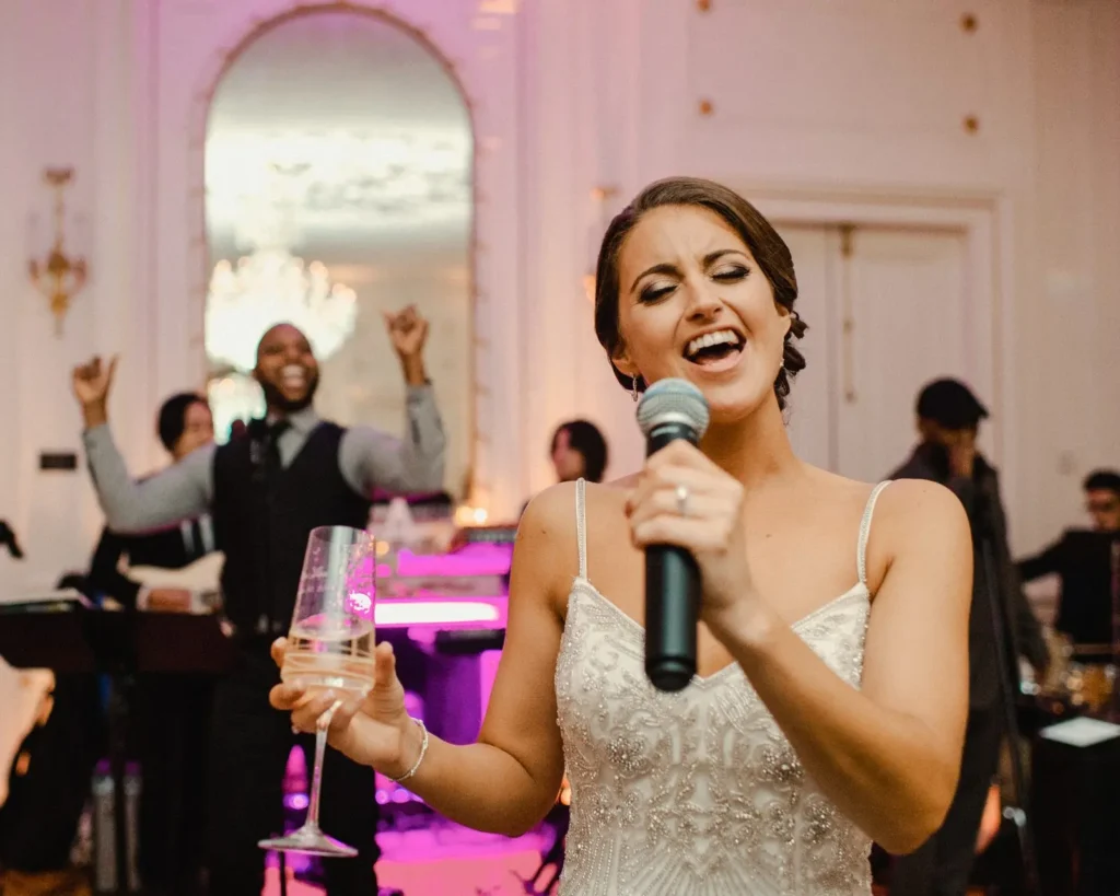 Bride sings into a microphone with guests cheering behind her at a Tupper Manor reception