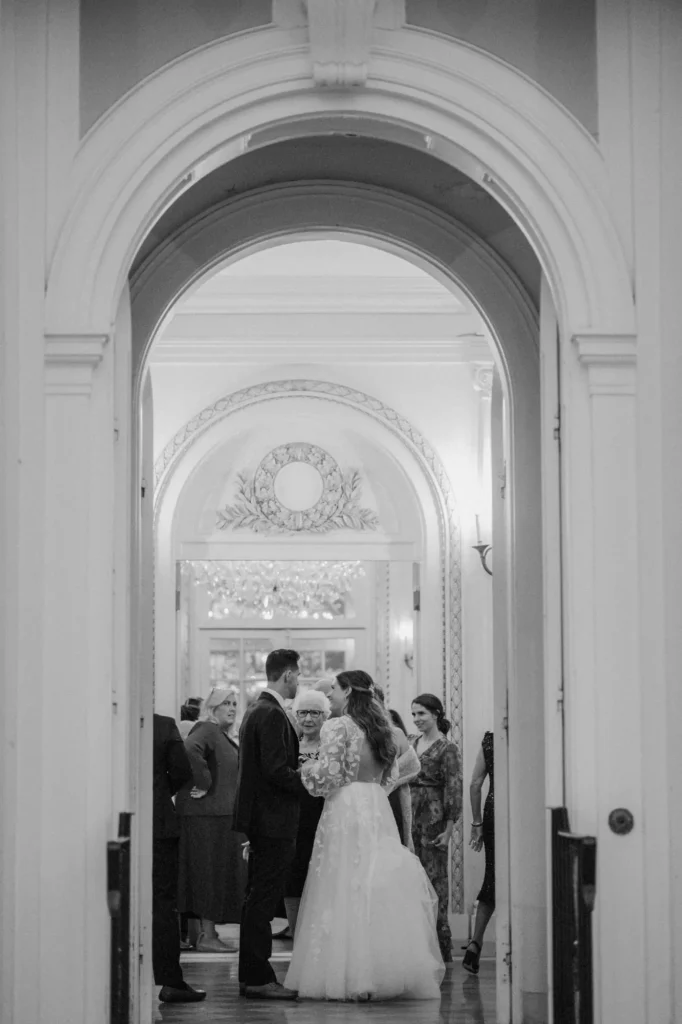 Black and white view through the arched doorway at Tupper Manor of the bride and groom greeting guests during cocktail hour