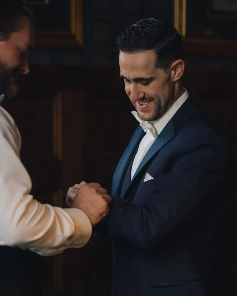 Groom laughs with a groomsman while getting ready in the wood-paneled room at Tupper Manor