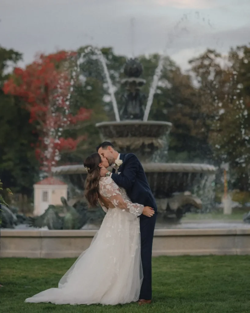 Bride and groom kiss in front of the fountain at Tupper Manor with fall foliage behind