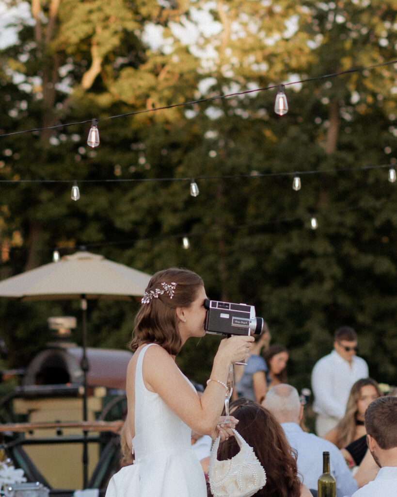 A bride filming her own reception with a vintage movie camera, string lights and guests behind her